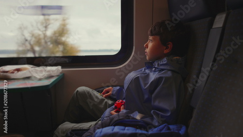 Photography Young boy sits alone on train seat holding small toy while watching scenery pass