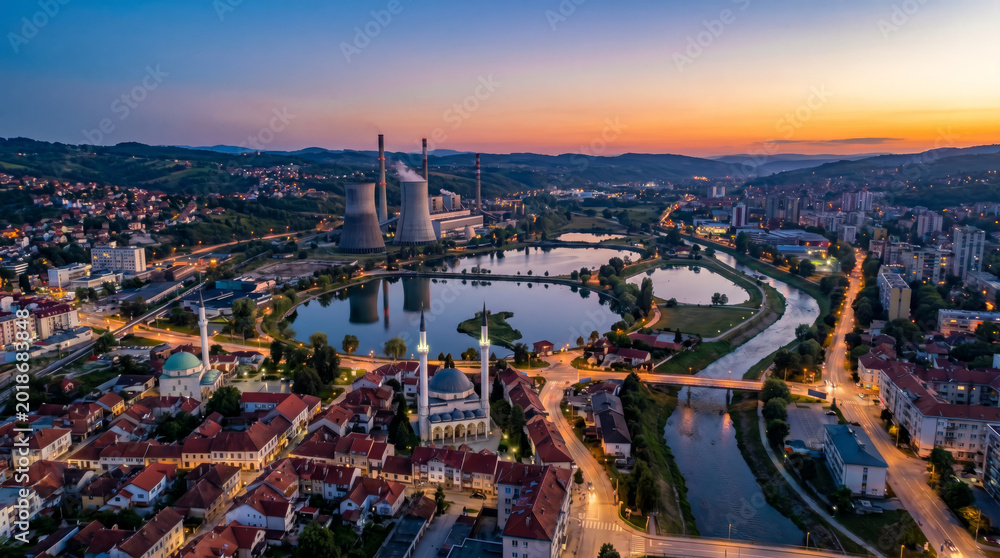 Fototapeta premium Aerial view of a city with a power plant and a river at sunset, showcasing urban development and industrial infrastructure.
