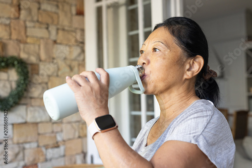 Senior Asian woman drinking from blue bottle wearing square smartwatch near French doors at home