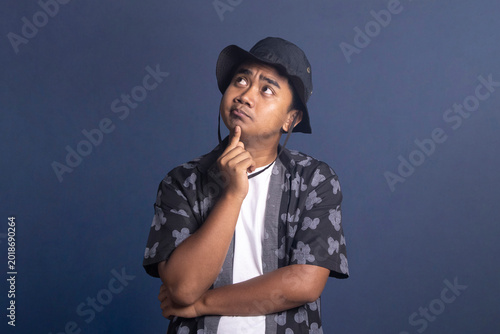 Bild auf Leinwand Young man wearing a black bucket hat and patterned shirt looking upward with a thoughtful and pensive expression while touching his chin