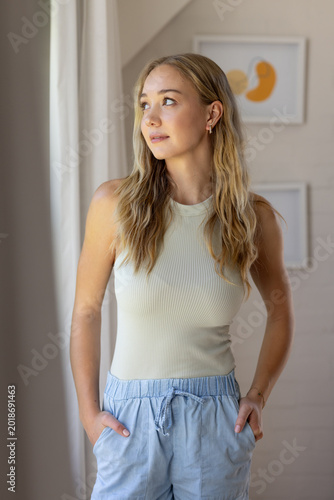 Adult female posing by window at home, gazing at sheer curtain wearing ribbed top, drawstring pants