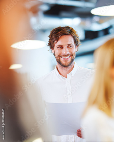 A smiling man with curly hair and a white shirt engages in a friendly conversation in a bright, modern office space.