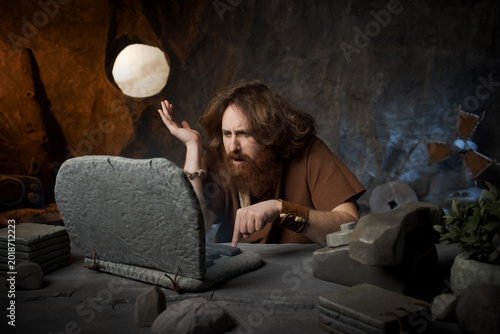 Confused caveman looking at a system error on his stone laptop screen with a frustrated expression.