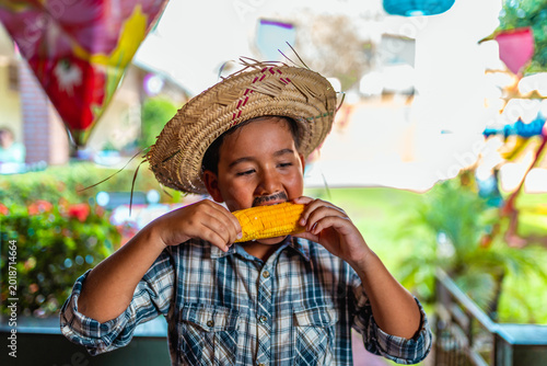 Young boy in traditional straw hat and plaid shirt eating corn on the cob during a Brazilian Festa Junina celebration.