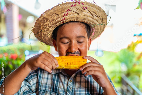 Young boy in traditional Festa Junina costume with straw hat and fake mustache eating corn on the cob.