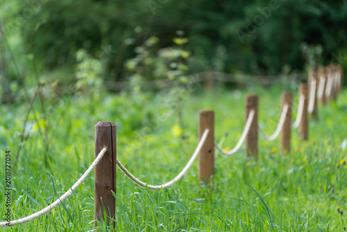 Rustic rope fence lines a vibrant green meadow. Wooden posts support thick, natural rope barriers. Background shows soft-focused forest and wildflowers. Ideal for nature, travel