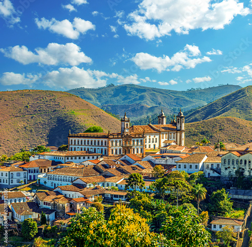Fotografie Panoramic view of the historic city of Ouro Preto, Minas Gerais, Brazil, featuring colonial architecture, Baroque churches, and terracotta roofs nestled against lush green mountains under a blue sky