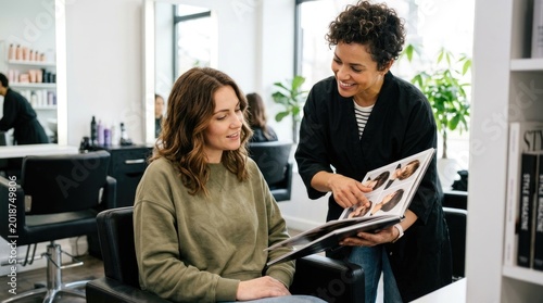 A stylist is showing a client a photo album of hairstyles. The woman is sitting in a salon chair, and the stylist is standing beside her, pointing at the images.
