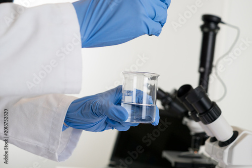 Valokuva Detailed view of a scientist's gloved hands carefully adding a concentrated blue substance into a beaker filled with water, symbolizing chemical reactions or viral staining during research