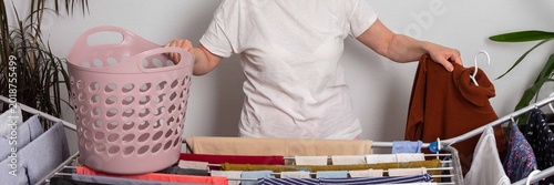 A 50-year-old woman with glasses hangs washed clothes on a drying rack in her indoor space. She sorts through the laundry from a pink basket as she prepares it to dry, banner