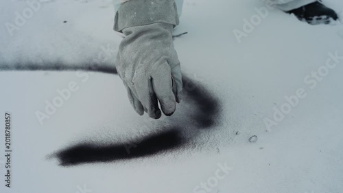 Hand sprinkles black powder on snow to create Sshaped trail, gloved technician draws sweeping curve, fine grains scatter across pristine surface, boot prints nearby, closeup contrast between dark