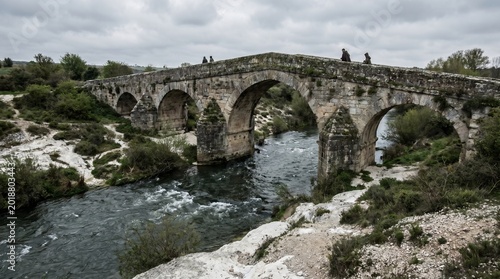 An old stone bridge with multiple arches spans a river on a cloudy day