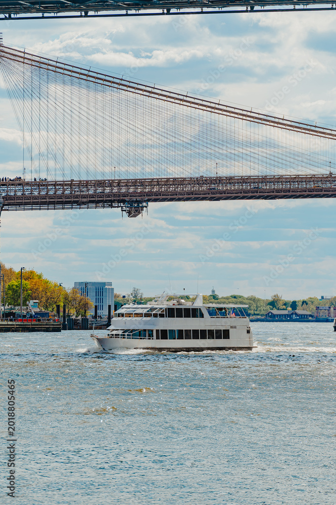 Obraz premium White Ferry Passing Beneath Brooklyn Bridge In New York. White passenger ferry crosses East River beneath Brooklyn Bridge cables in New York, daytime.