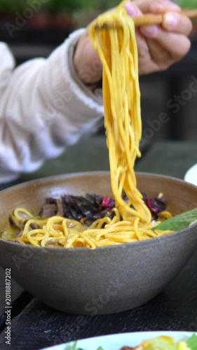 Close up of a person lifting delicious yellow ramen noodles with wooden chopsticks from a dark bowl. A fresh salad is visible in the background. Showing a healthy and appetizing meal at a restaurant