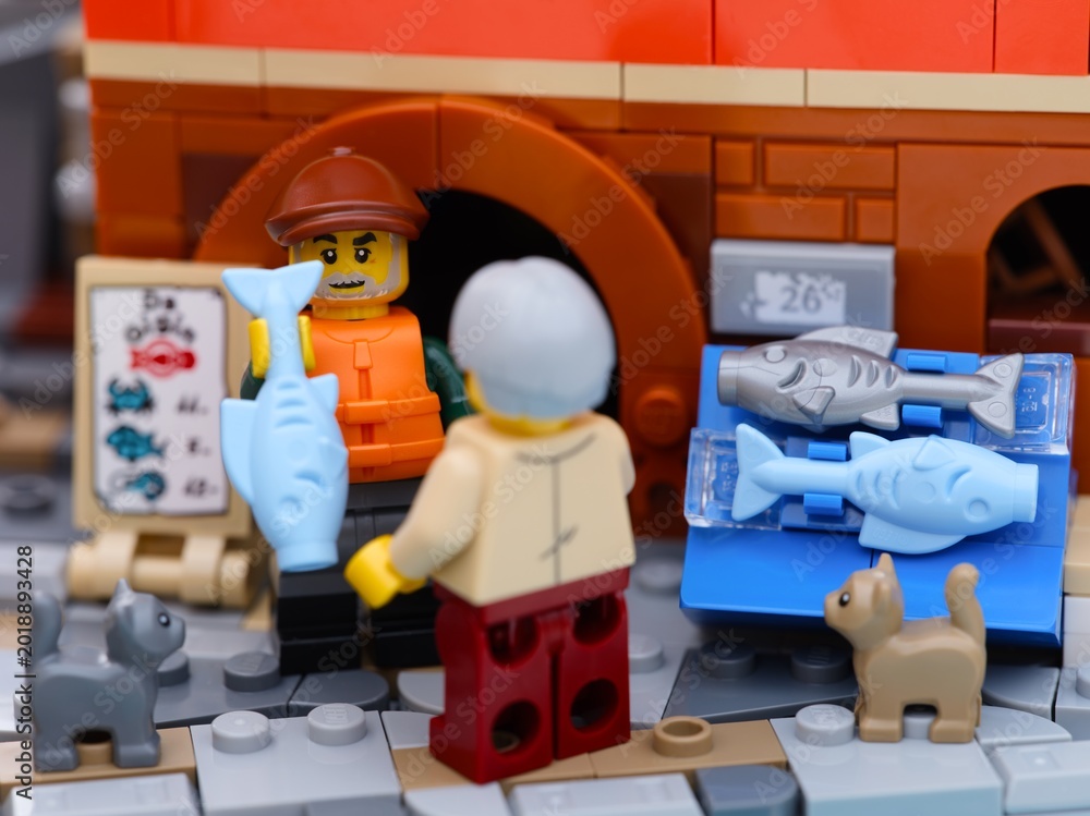 Obraz premium Tambov, Russian Federation - January 19, 2026 Lego fisherman minifigure selling a light blue fish from a fish stall to a customer on a city street. Two Lego cats standing near.