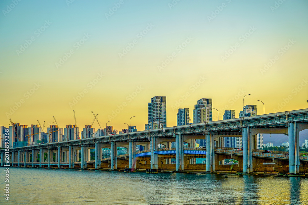 Fototapeta premium Han River Expressway Bridge at Warm Sunset with Active Construction Cranes and Modern Apartment Buildings Rising Behind, Seoul South Korea