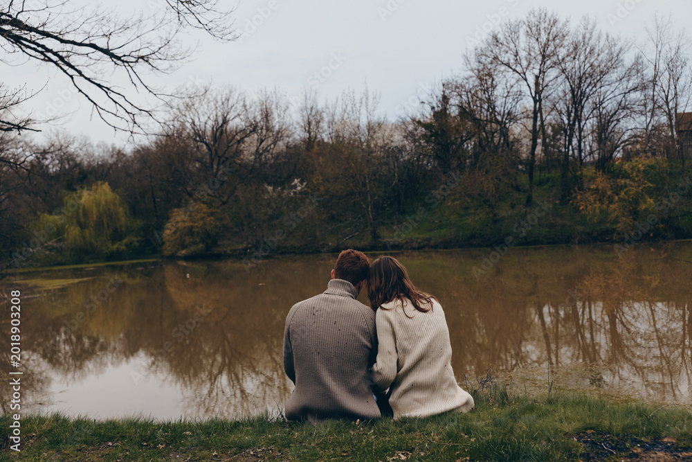 Naklejka premium A couple sits close by a calm lake in autumn, wearing cozy sweaters, sharing a quiet moment, soft breeze, muted colors, reflective water, peaceful countryside setting