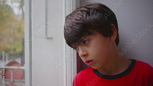 Obraz na plátne Boy standing quietly by window with thoughtful expression while gazing outside c