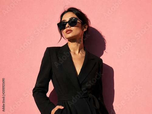 Elegant woman in black blazer and sunglasses posing against a pink wall.