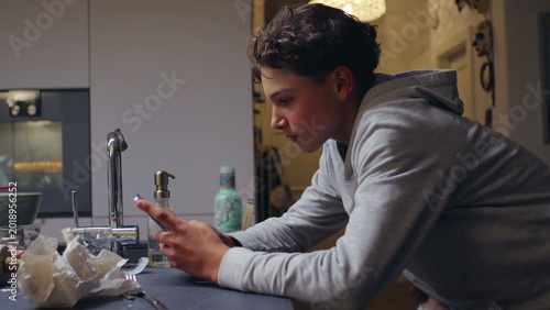 Teenager leaning over kitchen counter using smartphone with focused expression, absorbed in quiet digital moment inside home environment, conveying modern contemplative lifestyle