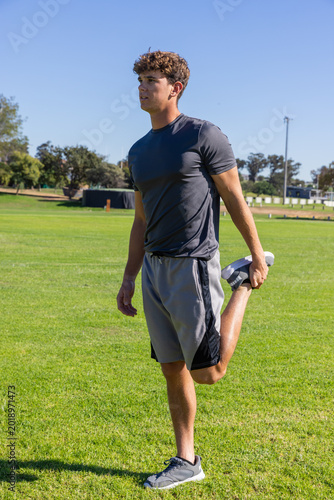 Fotografie Male performing standing quadriceps stretch on grass athletic field wearing runn
