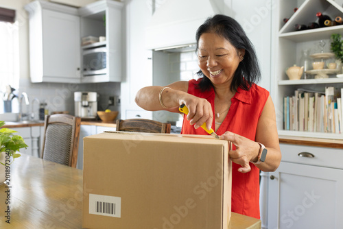 Senior Asian woman in red blouse standing at kitchen dining table opening box with yellow cutter