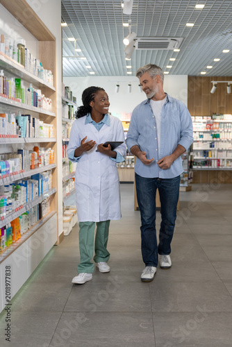 African American female pharmacist with tablet talking and walking with Caucasian man between shelves