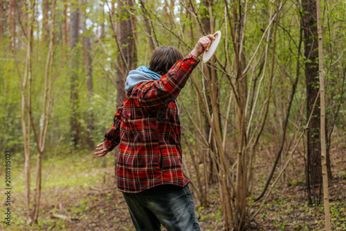 Fotografi Male wearing red plaid shirt and blue hoodie throws a frisbee in a green forest,