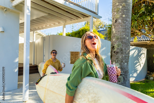 Obraz na plátně Happy women surfers carrying boards laughing in front of beach house