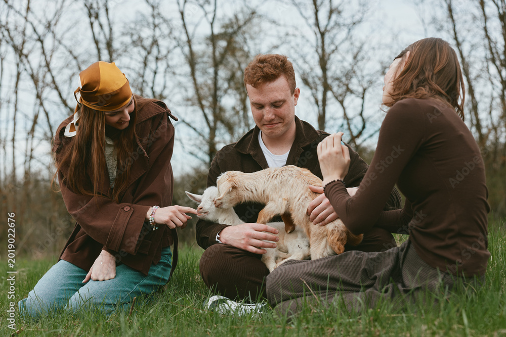 Naklejka premium Three people share a playful moment with a goat kid in a grassy meadow, outdoors lifestyle scene. They laugh and interact, showcasing bonding, care, and rustic countryside vibes in natural light.