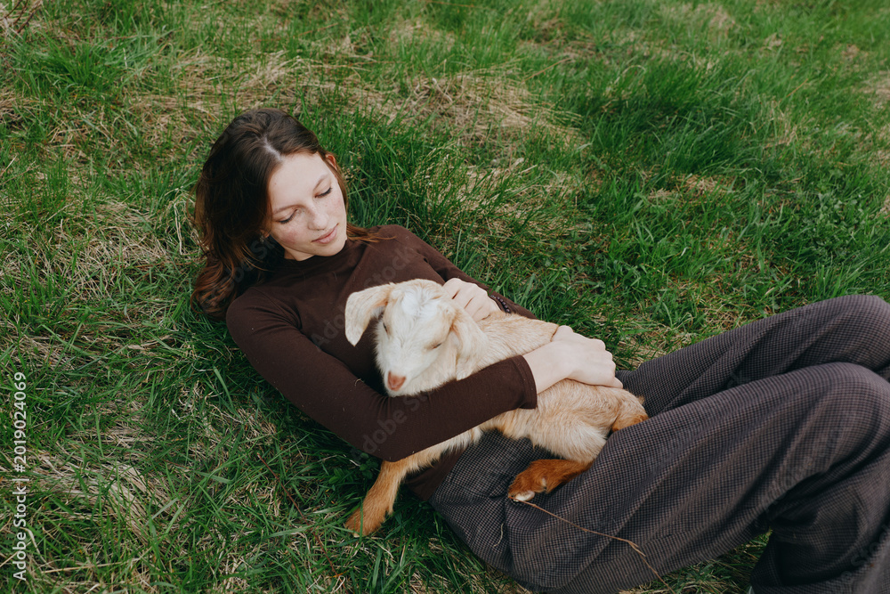 Naklejka premium Outdoor scene of a woman lying on lush grass, gently cuddling a goat kid, sun warmed, cozy and relaxed, showcasing lifestyle, companionship, and a deep connection with nature
