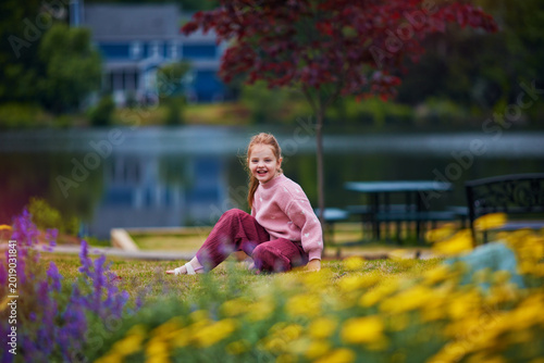 Smiling Little Girl Sitting by the Lake Surrounded by Blooming Flowers in Park - Playful Summer Childhood Lifestyle