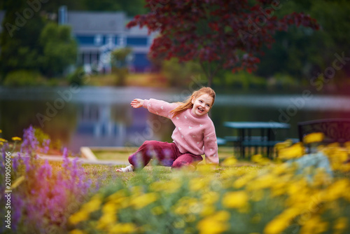 Smiling Little Girl Sitting by the Lake Surrounded by Blooming Flowers in Park - Playful Summer Childhood Lifestyle