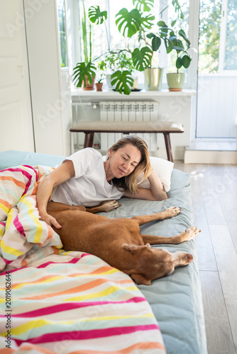 Foto Relaxed woman gently stroking Magyar Vizsla, lounging on bed in morning