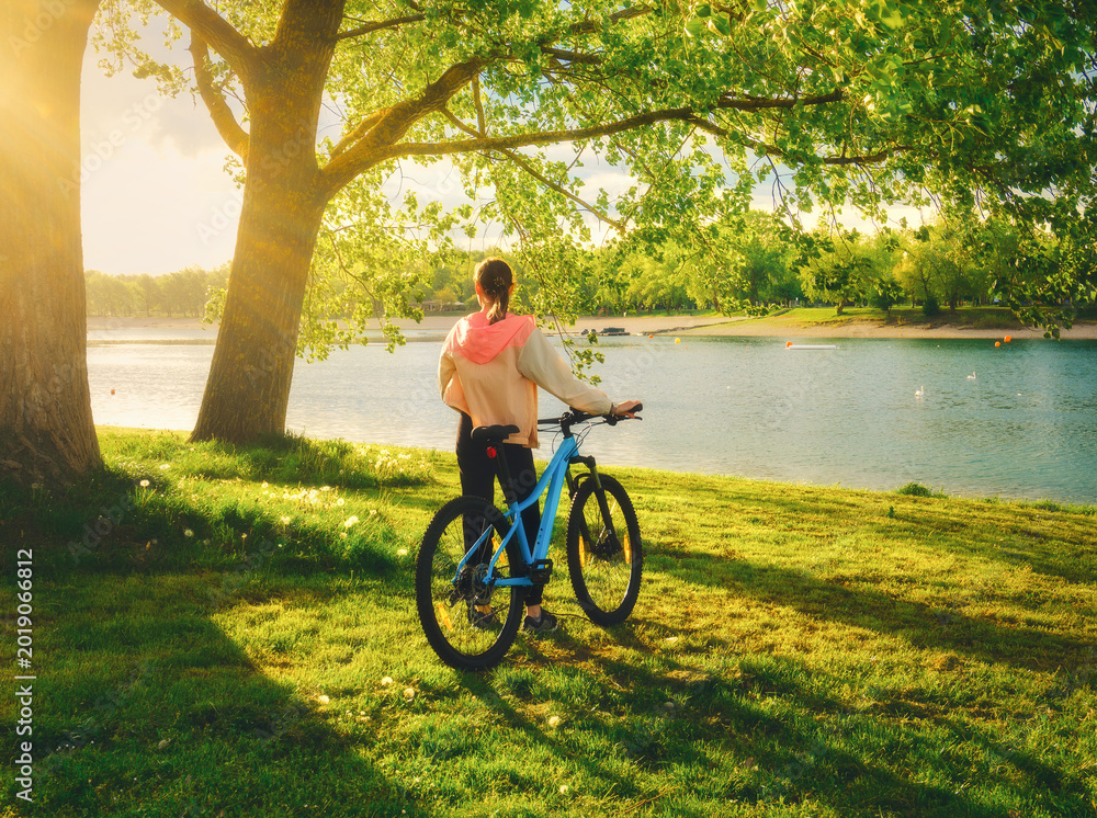 Obraz premium Woman riding a mountain bike near green trees and lake at sunset in spring. Colorful landscape with sporty girl, bicycle, beach, river, green grass, river in park. Summer. Sport and travel. Biking