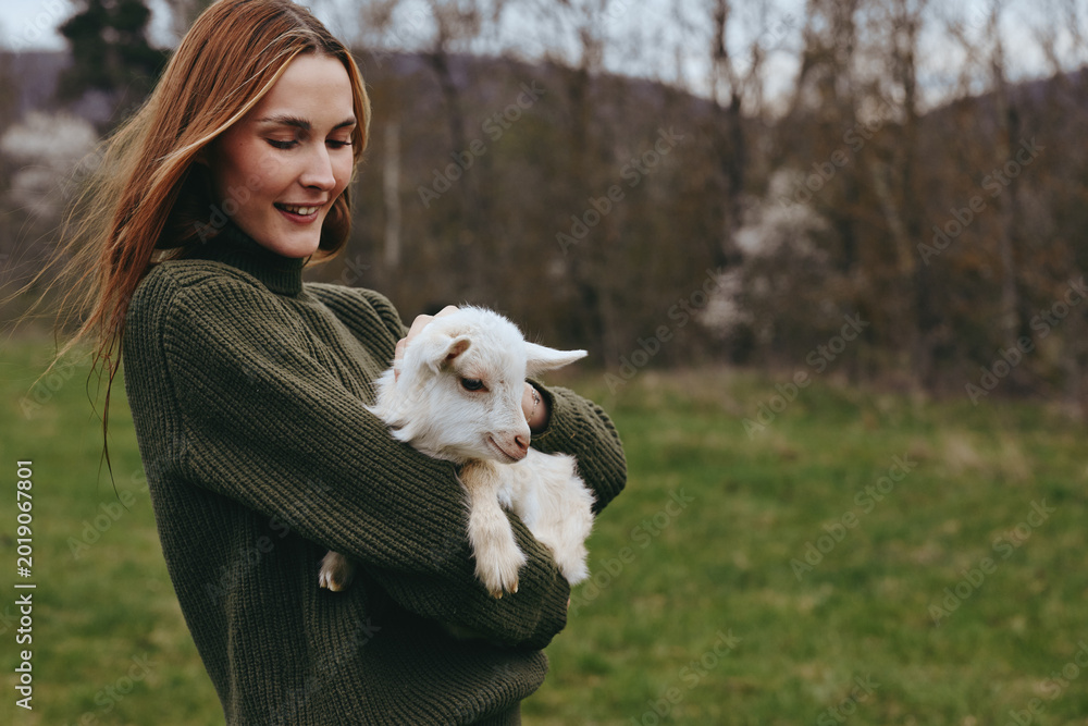 Obraz premium A woman in a dark green sweater holds a fluffy white goat kid in a green field, presenting a warm countryside lifestyle moment with gentle touch and peaceful animal companionship