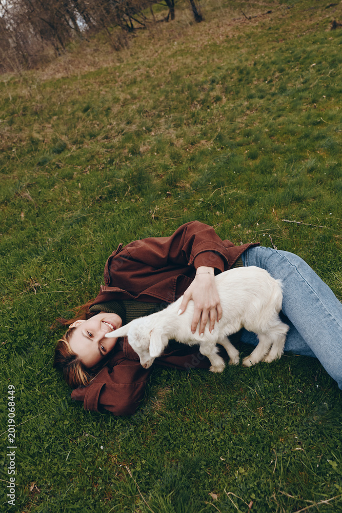 Obraz premium Young person lying on grass with playful goat kid, enjoying a relaxed outdoor moment, casual clothing, gentle interaction and affection in a tranquil park