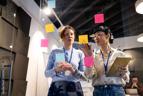 Diverse businesswomen collaborating efficiently, brainstorming ideas using sticky notes on glass wall and digital tablet in modern office.
