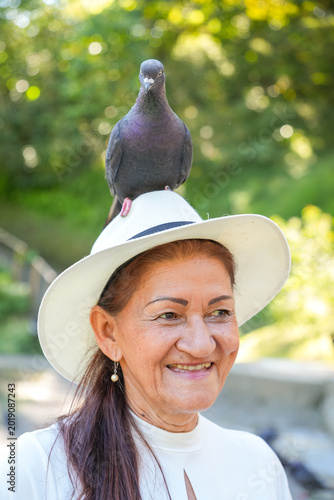 An elderly woman wearing a hat with a dove in her head smiles peacefully while enjoying a sunny day outdoors in a green park