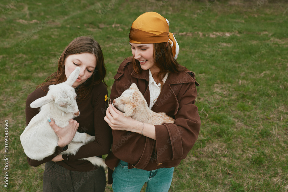 Naklejka premium A pair of women share a gentle moment with a baby goat in an open grassy field, capturing a wholesome lifestyle scene focused on animal care, friendship, and outdoor farm life.