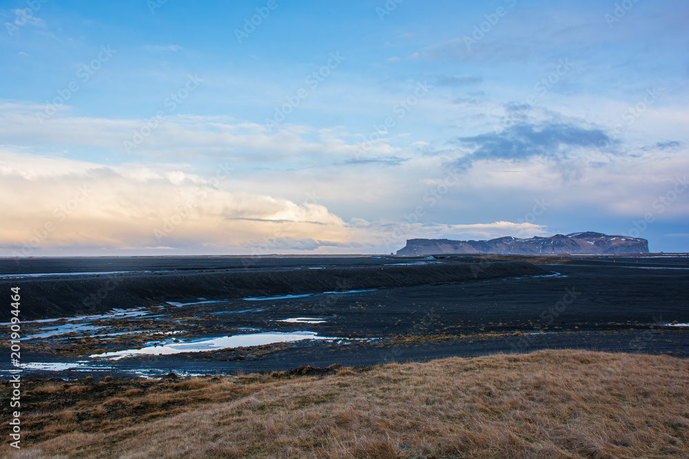 Fototapeta premium Vast volcanic landscape with black sand plains and distant mountains under a cloudy sky in Iceland. High quality photo
