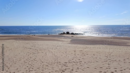 A wide angle view of Praia de Miramar in Portugal shows footprints on soft sand and rocky outcrops in the water. Bright sunlight creates a shimmering reflection on the calm blue Atlantic Ocean.