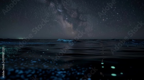 Night sky with glowing bioluminescent waves on ocean water under stars.