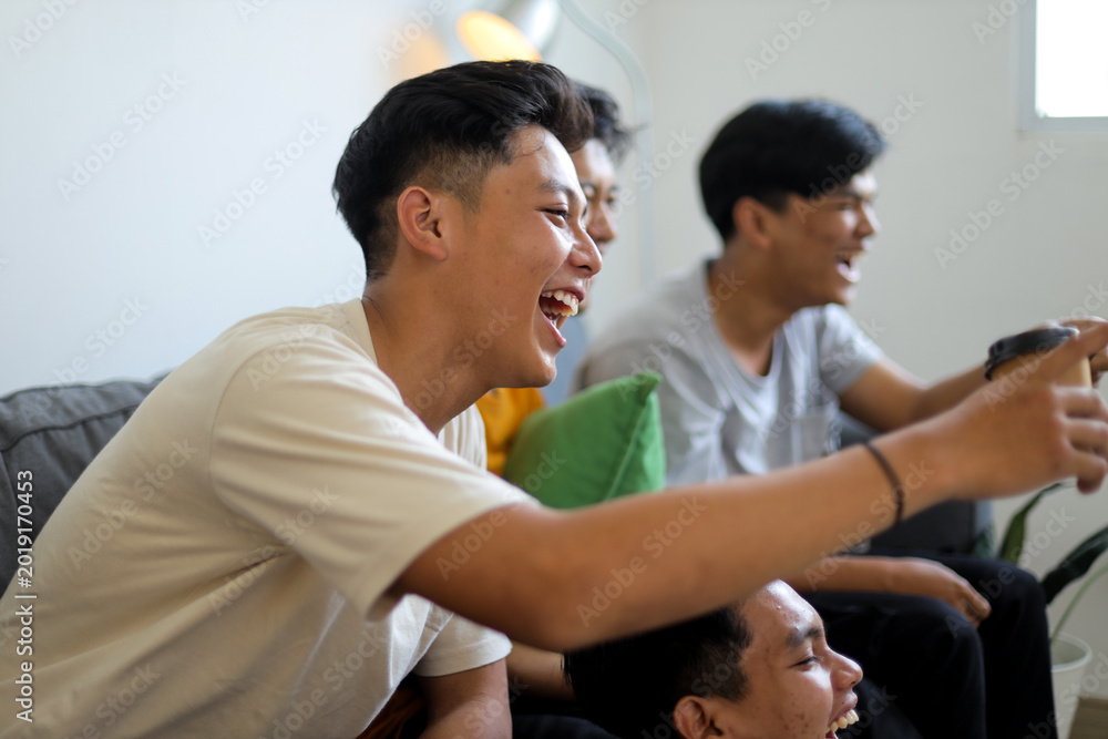 Fototapeta premium Group of Cheerful Asian Young Men Sitting on Sofa, Laughing and Enjoying Gathering in Living Room