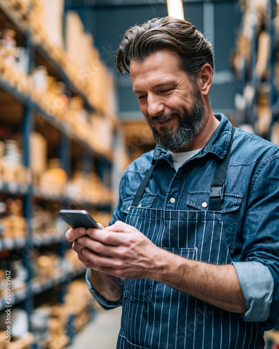 Handsome Business Owner in Warehouse  Bearded Man in Blue Shirt Using Smartphone for Inventory Management