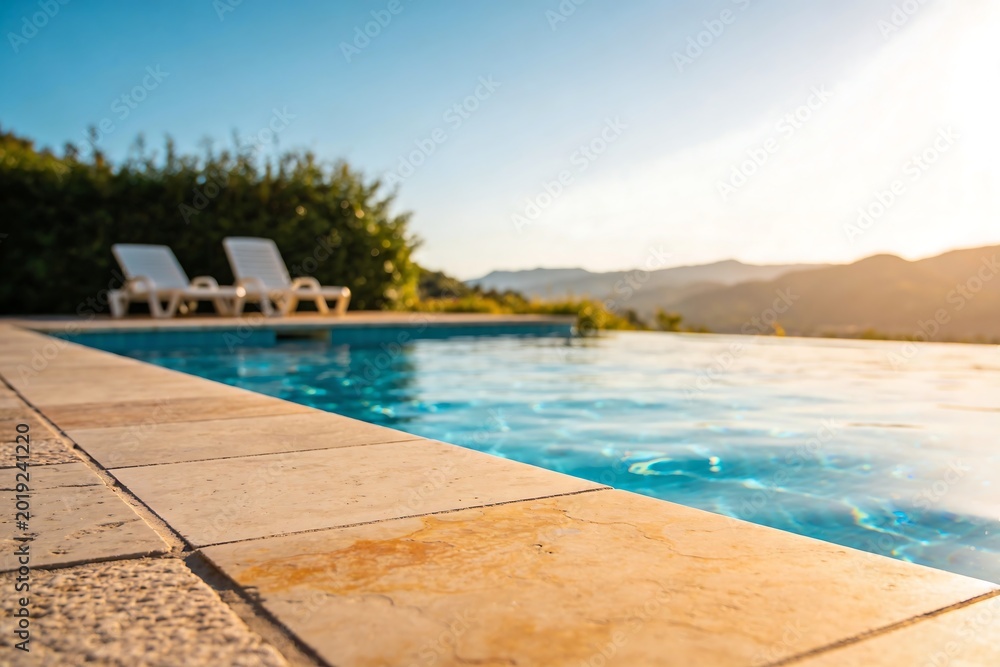Fototapeta premium Modern luxury villa poolside with travertine tiles and clear blue water. Sunlit terrace with lounge chair, surrounded by tropical nature, perfect vacation relaxation lifestyle scene.