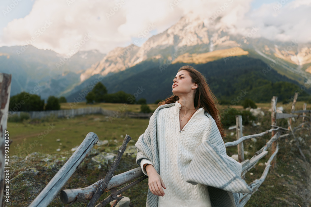 Fototapeta premium Woman in a cozy poncho stands beside a rustic fence in a mountain landscape, enjoying fresh air and a quiet moment, embracing nature during outdoor lifestyle.
