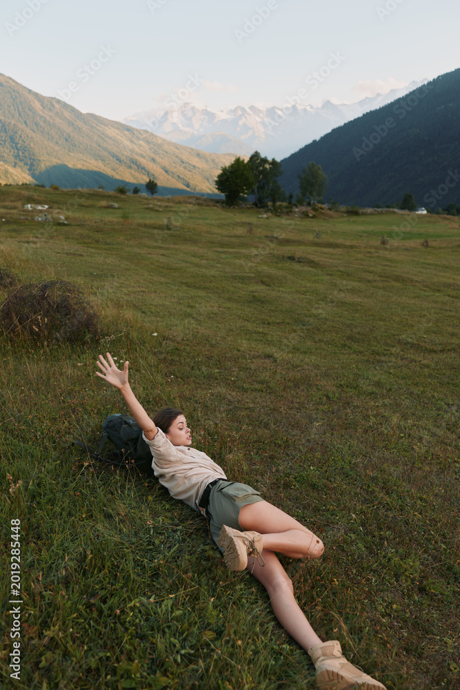 Fototapeta premium Relaxed woman lying on a grassy meadow with distant mountains, enjoying sunshine and fresh air, captured as a casual outdoor lifestyle moment in a natural landscape setting.