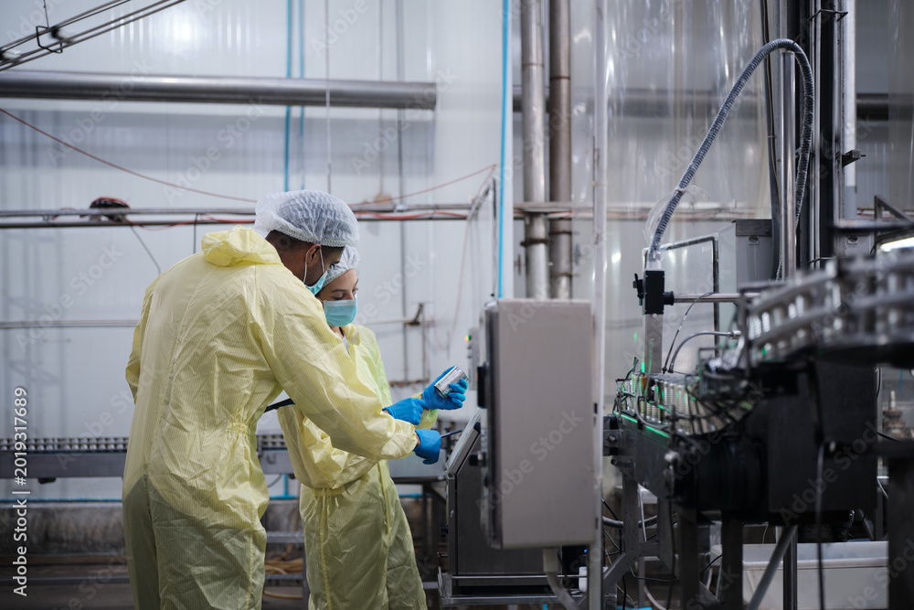 Fototapeta premium Two industrial engineer supervisors examine quality of steel containers on factory conveyor belt, inspect production process in canned food manufacture, and protective uniforms for hygiene operations.