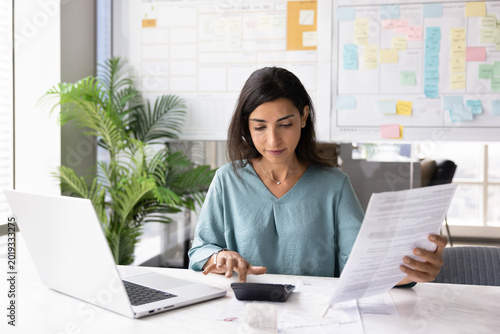 Focused professional woman reviewing financial documents, holds invoice, preparing report or budget summary, using calculator for expenses and incomes calculations seated at workplace desk with laptop